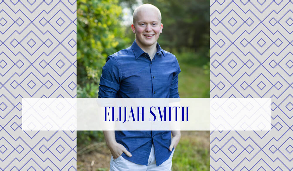 A man stands outdoors in casual attire, wearing a blue button-up shirt and light-colored pants, smiling gently with one hand in his pocket. His clean-shaven head and fair complexion contrast with a leafy green backdrop, and a semi-transparent panel.